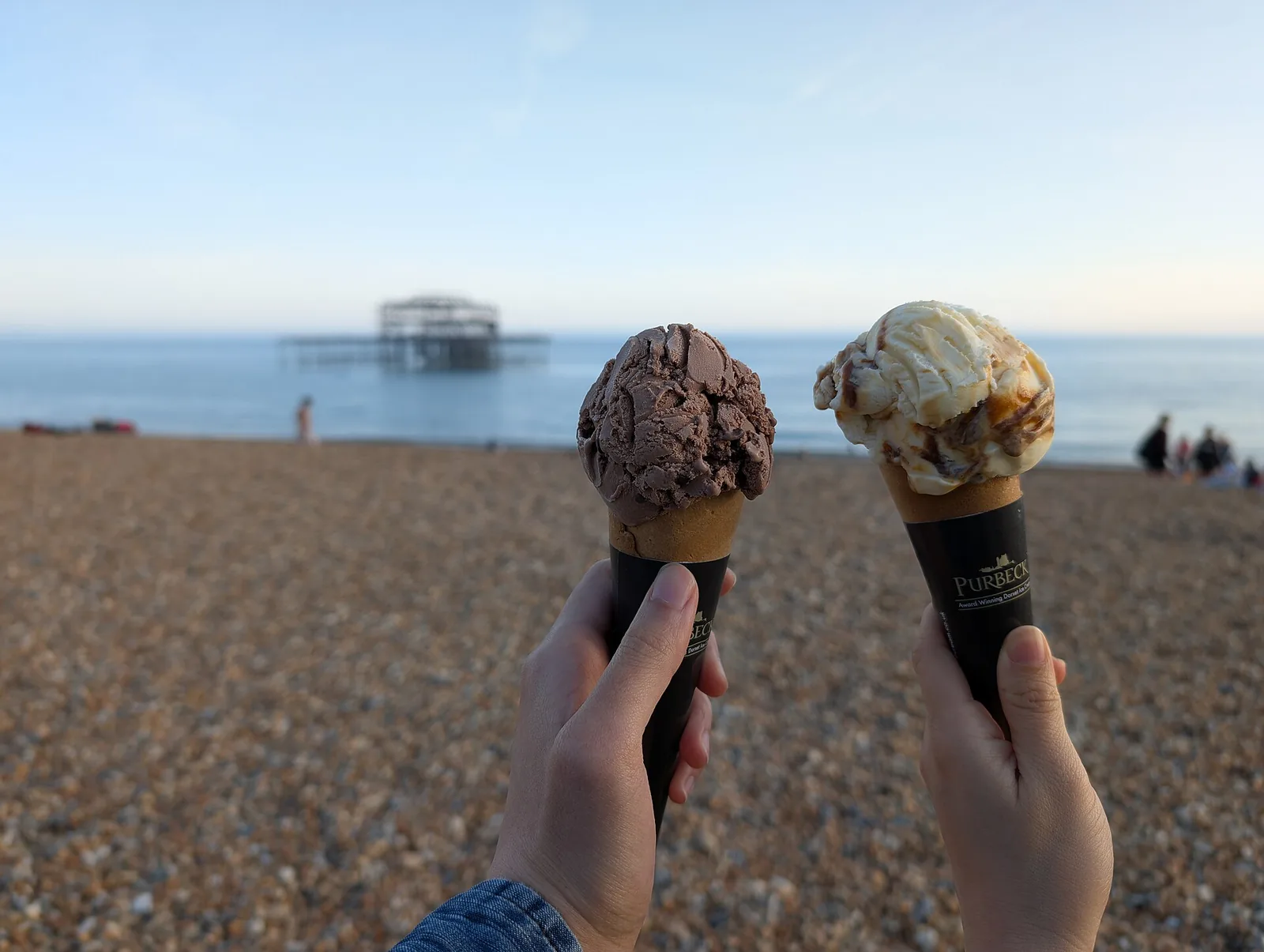 Two scoops of ice cream in cones, held up on a pebble beach.